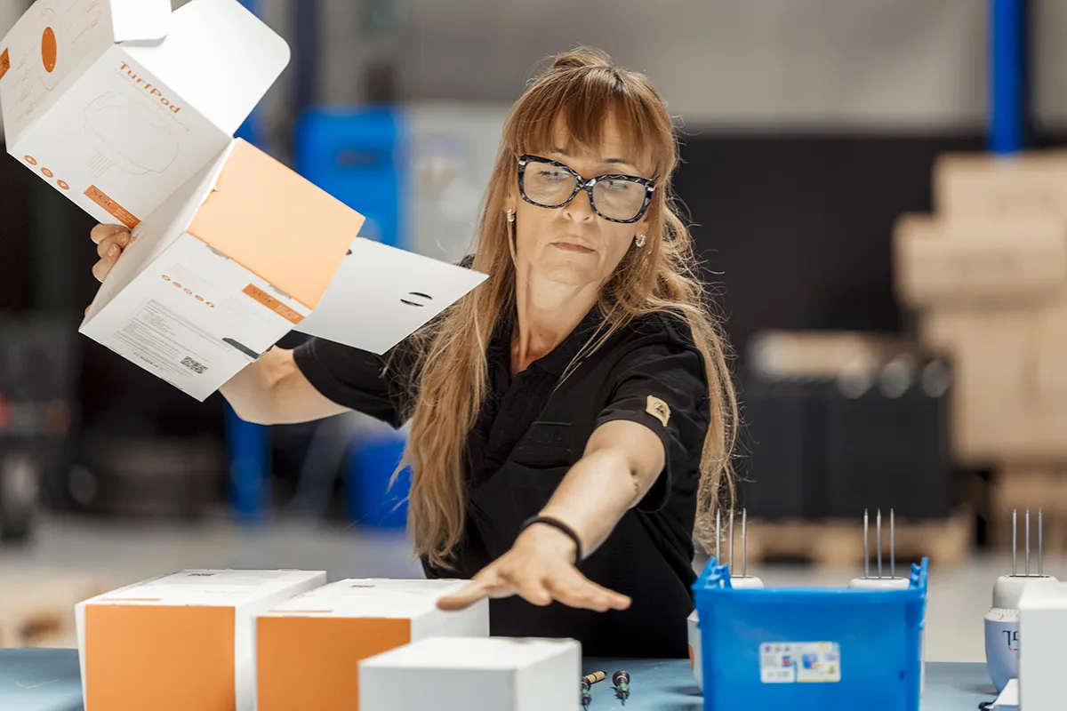 Een vrouw met lang bruin haar en een bril zet kartonnen dozen in elkaar op een werkplek. Ze strekt haar hand uit om een blauw onderdeel, dat lijkt op een elektronisch apparaat, op tafel te plaatsen. De achtergrond is een wazige industriële omgeving met verschillende apparatuur en dozen zichtbaar.