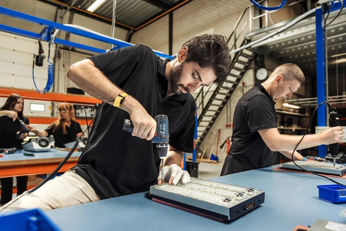 Twee mannen in zwarte shirts werken op een bank in een fabriek aan het assembleren van elektronische componenten. Ze gebruiken gereedschap onder fel licht. Op de achtergrond zijn drie andere arbeiders te zien die soortgelijke taken uitvoeren. Er zijn planken met apparatuur en een trap zichtbaar, wat wijst op een industriële setting.