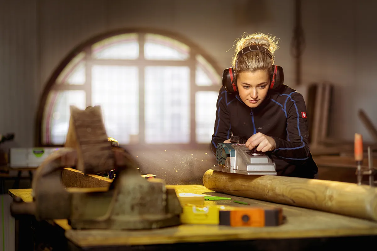 Een vrouw, gekleed in een blauw-zwart jasje en een rode hoofdtelefoon met ruisonderdrukking, bedient een schaafmachine op een houten balk in een timmerwerkplaats. Zaagsel is zichtbaar en er zijn verschillende gereedschappen, waaronder een waterpas en een bankschroef, op de werkbank geplaatst. Een groot, gebogen raam op de achtergrond zorgt voor natuurlijk licht.