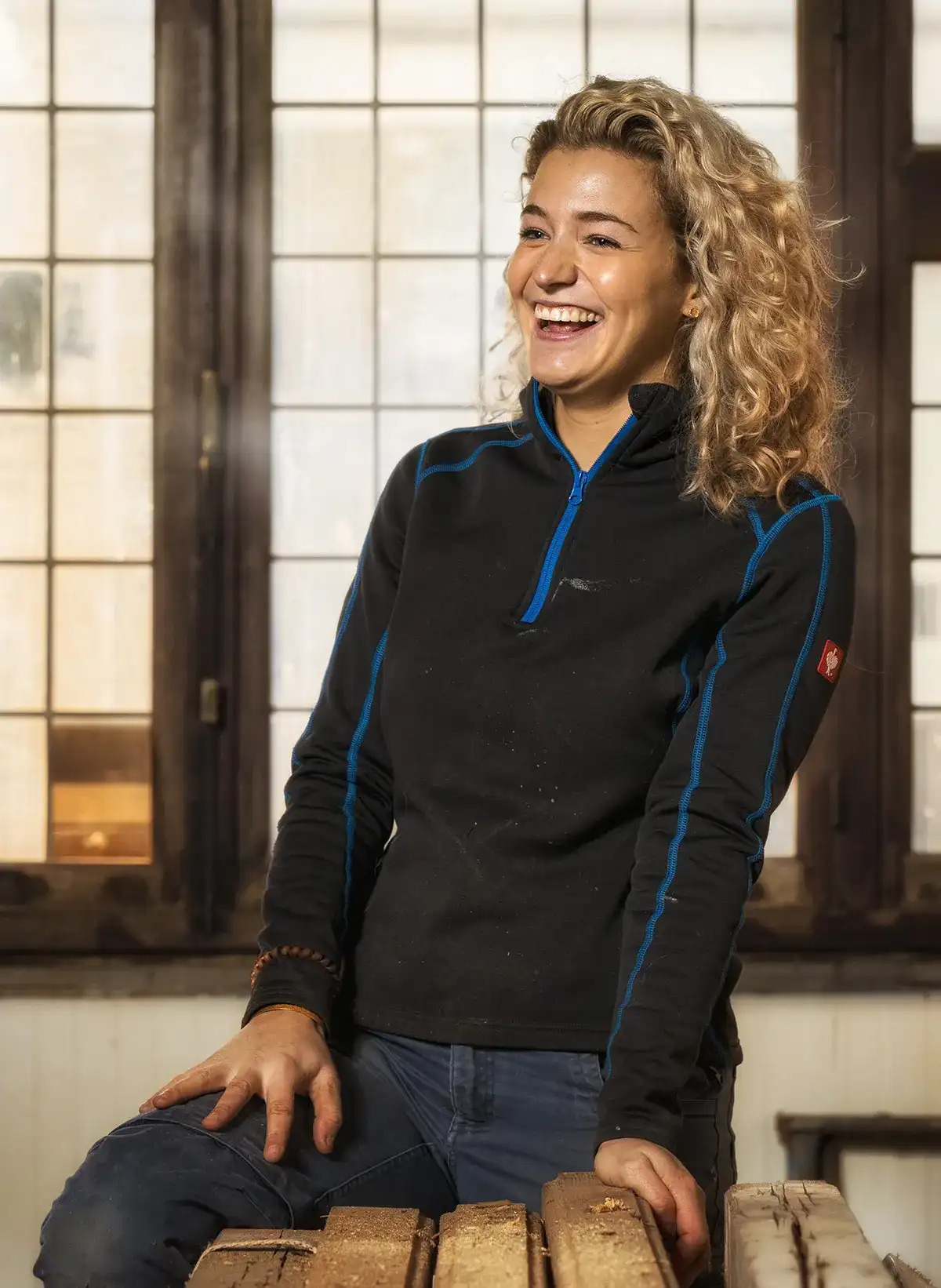 Een vrouw met blond krullend haar staat binnen voor een groot raam met rasterachtige ruiten. Ze draagt een zwart topje met lange mouwen en blauwe accenten en glimlacht breed terwijl ze op een stapel houten planken leunt. De achtergrond is warm verlicht, waardoor er een gezellige sfeer ontstaat.