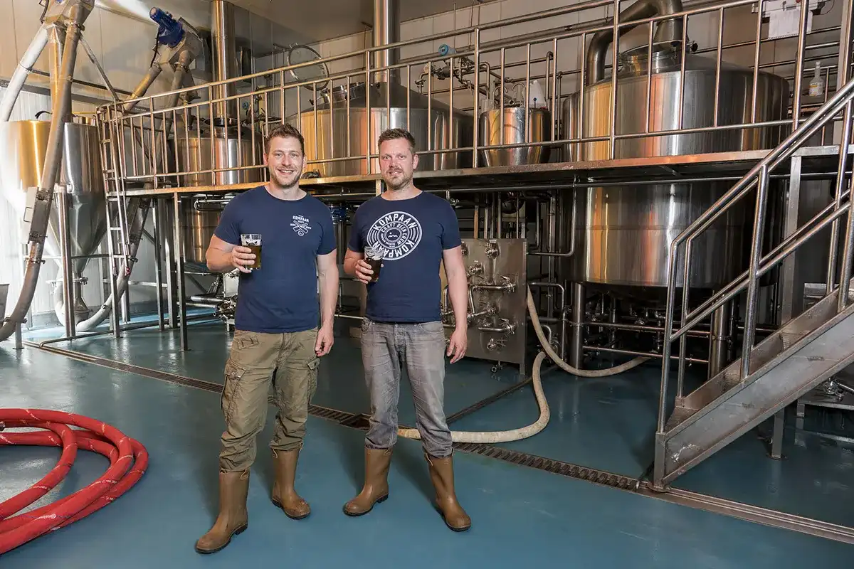 Twee mannen, allebei gekleed in marineblauwe T-shirts en bruine laarzen, staan in een brouwerij. Ze houden elk een glas bier vast en glimlachen naar de camera. Daarachter is industriële brouwapparatuur zichtbaar, waaronder grote roestvrijstalen tanks en diverse leidingen. De vloer is blauw en aan de linkerkant liggen rode slangen.