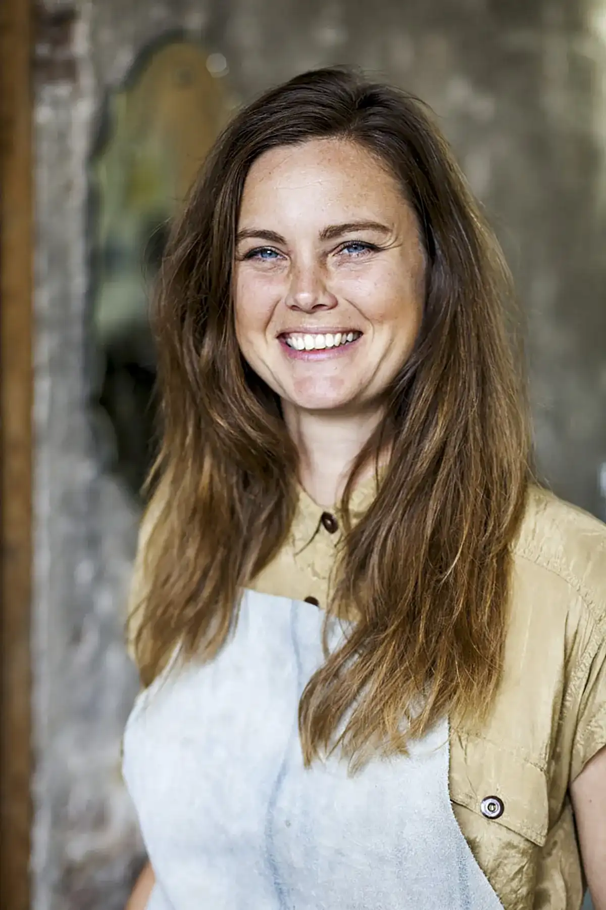 Een lachende vrouw met lang bruin haar staat binnen. Ze draagt een beige overhemd met knoopjes en een lichtblauw schort. De achtergrond is enigszins vervaagd en toont een rustieke omgeving met een mix van texturen en kleuren.