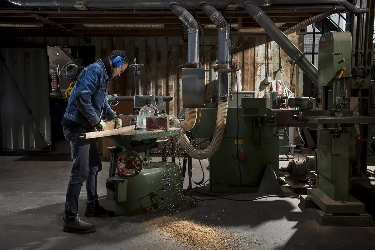Een persoon die beschermende oorkappen en een bril draagt, bedient een grote groene houtbewerkingsmachine in een werkplaats. Ze voeren een houten plank in de machine, waardoor er zaagsel op de vloer ontstaat. De werkplaats is industrieel met diverse andere machines en apparatuur op de achtergrond.
