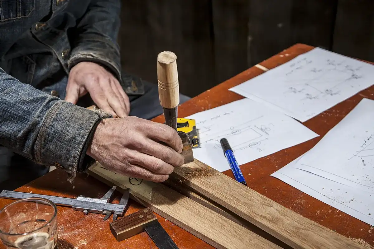 Een persoon gebruikt een beitel om een stuk hout te snijden. De werkbank ligt bezaaid met houtbewerkingsgereedschap, blauwdrukken en papieren. Een deel van de persoon is zichtbaar en draagt een spijkerjasje. De scène is goed verlicht, waarbij de nadruk ligt op het gedetailleerde werk dat aan het houten object wordt gedaan.