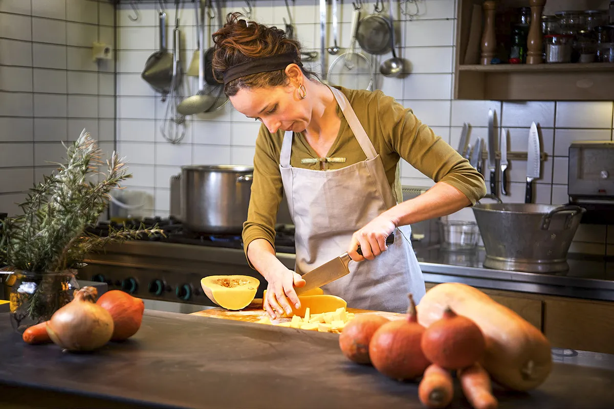 Een vrouw, gekleed in schort en hoofdband, staat in een keuken een pompoen te hakken. Op de toonbank staan diverse groenten, waaronder uien en nog meer pompoenen. De keuken heeft betegelde muren, potten, pannen en keukengerei hangen op de achtergrond, en een grote pot op het fornuis.