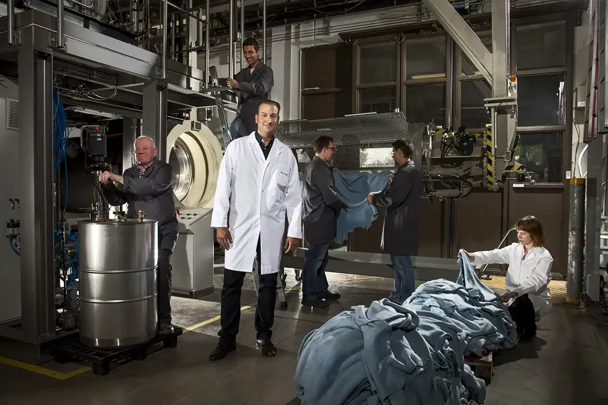 Een man in een witte laboratoriumjas staat glimlachend in een fabrieksomgeving. Om hem heen zijn arbeiders bezig met verschillende taken: de een bedient machines, een ander kijkt in een container, twee hanteren stof op een transportband en een vrouw sorteert een stapel blauw textiel op de vloer. Industriële apparatuur omringt hen.