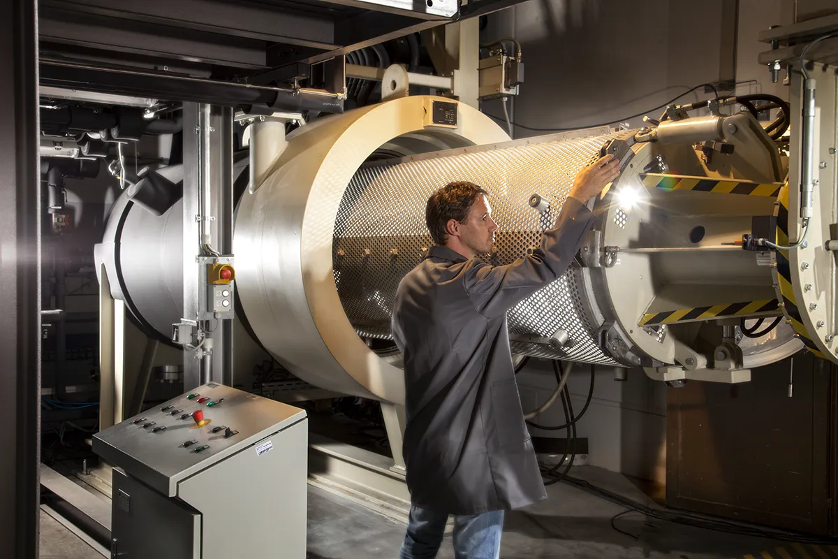 Een man in een zwarte laboratoriumjas bedient een grote, cilindervormige, hightech industriële machine met diverse bedieningspanelen en schakelaars. De achtergrond onthult een complexe opstelling van leidingen en machines in een goed verlicht laboratorium of productieomgeving. De scène benadrukt het gebruik van geavanceerde technologie.