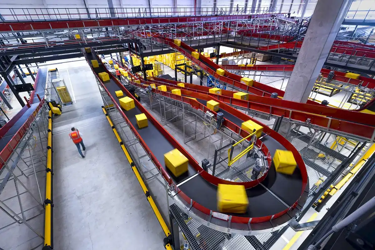 Een druk geautomatiseerd magazijn met meerdere rode transportbanden die talloze gele pakketten transporteren. Een arbeider in een oranje vest loopt langs de transportbanden. De industriële omgeving is helder verlicht met verschillende platforms, veiligheidsleuningen en zichtbare structurele balken.