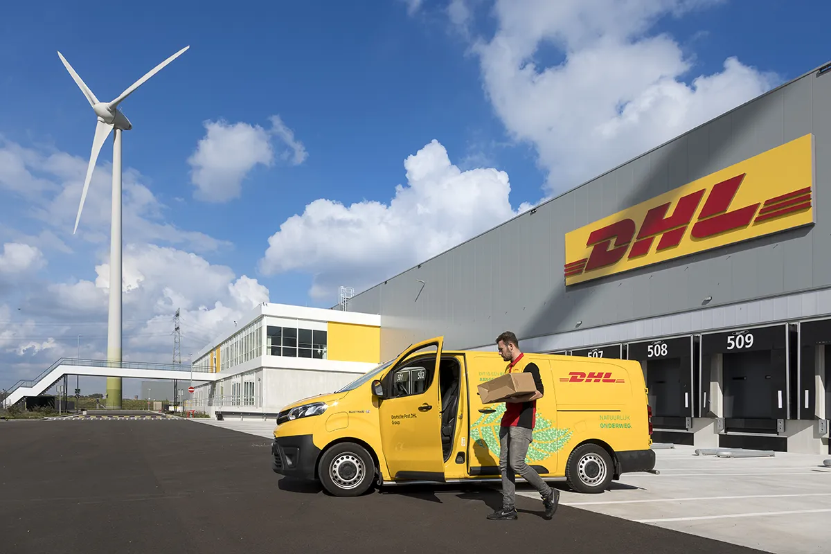 Een DHL-bestelwagen staat geparkeerd buiten een logistieke vestiging van DHL. Op zoek naar een bedrijfsfotograaf in Delft? Dan bent u bij Rogier Chang aan het juiste adres. Met fotoreportages voor onder meer de TU en andere bedrijven van de campus,op zijn naam, is hij regelmatig in Delft actief voor het maken van een ‘hi-tech’ bedrijfsreportage. Delft Een koerier in een grijs uniform met het DHL-logo draagt een pakketje weg van het busje. Het gebouw is modern en wit en het DHL-logo is prominent aanwezig. Op de achtergrond is er een grote windturbine en een gedeeltelijk bewolkte hemel.