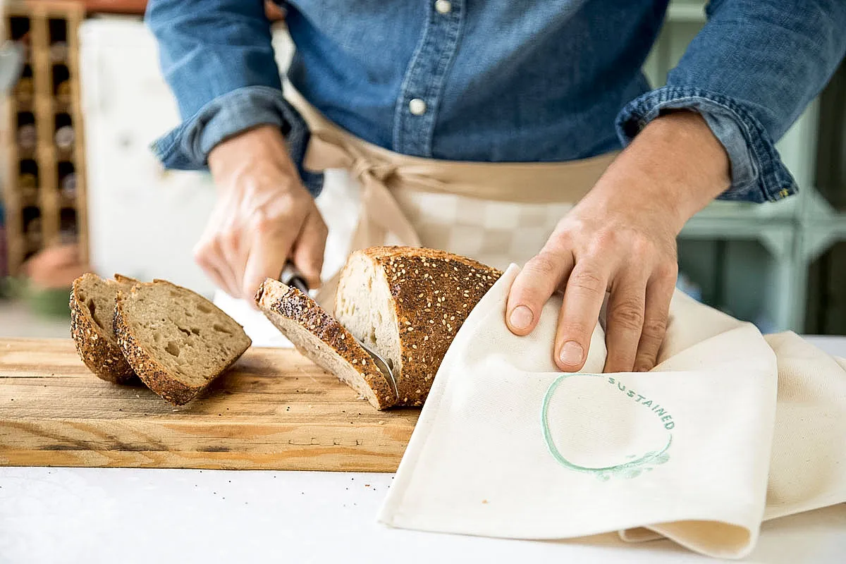 Een persoon die een spijkerblouse en een schort draagt, snijdt vers brood op een houten snijplank. Er zijn al twee sneetjes brood gesneden en opzij gelegd. Eén hand houdt het brood vast met een doek, terwijl de andere het mes geleidt. De achtergrond is enigszins wazig.