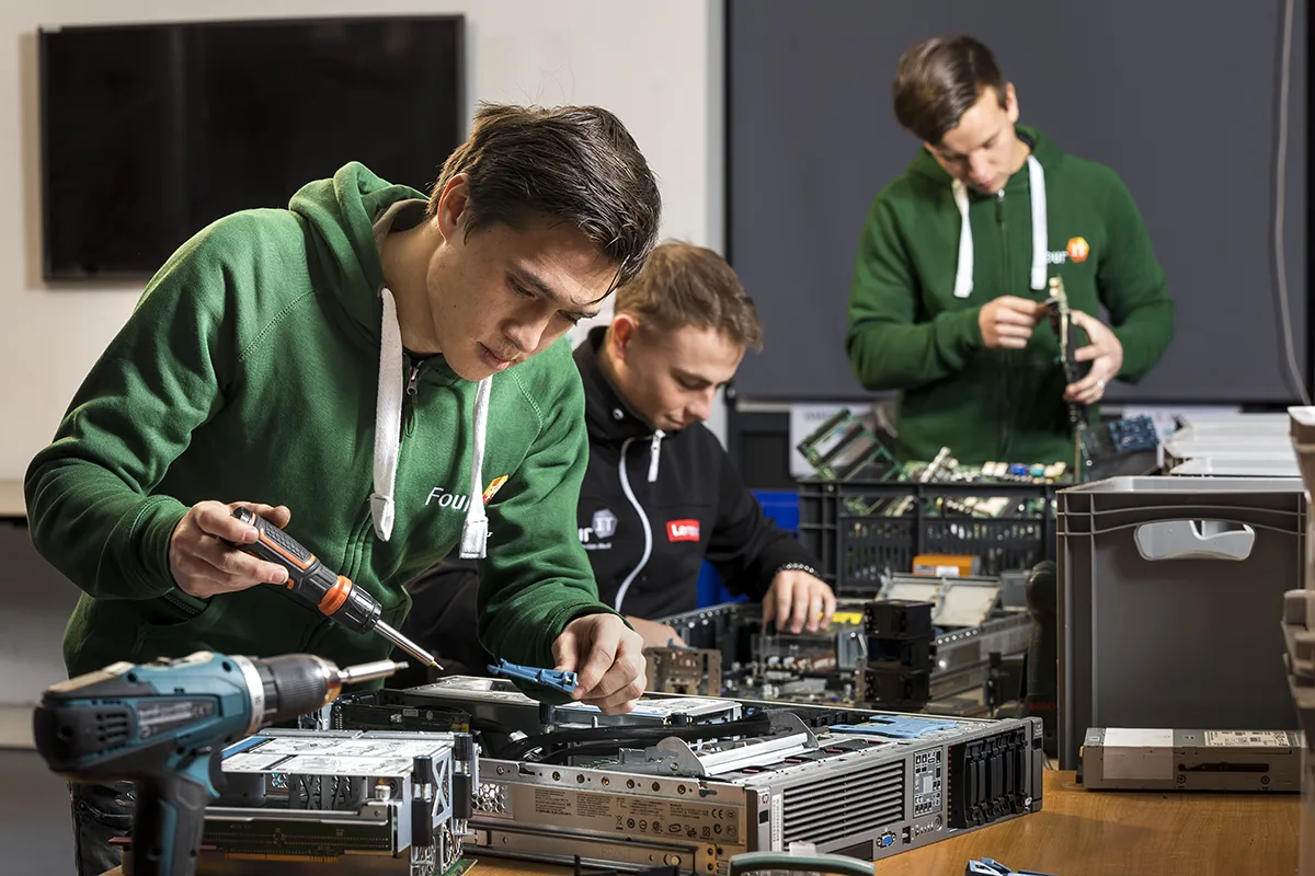 Drie mensen in groene hoodies zijn aan een tafel bezig met het in elkaar zetten van computeronderdelen. De één gebruikt een schroevendraaier op een desktoptoren, de tweede plaatst een onderdeel in een andere computer en de derde staat op de achtergrond een onderdeel te inspecteren. Diverse gereedschappen en elektronische onderdelen liggen verspreid.
