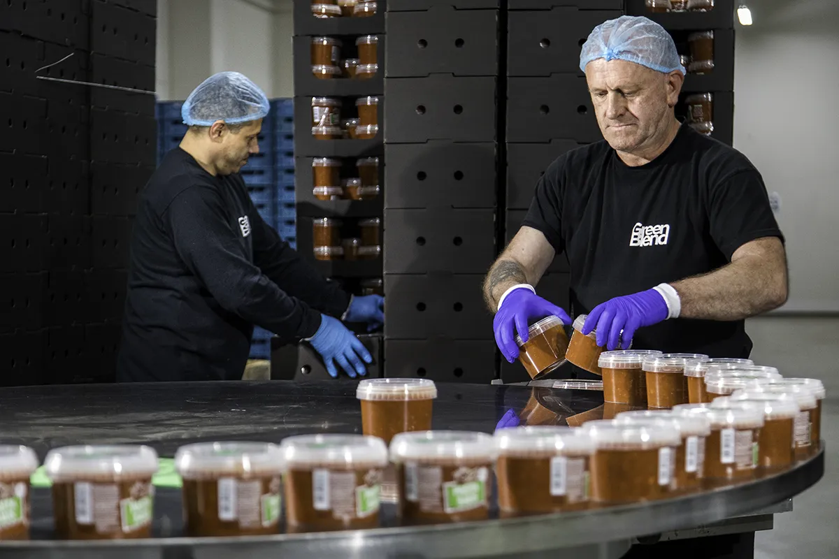 Twee fabrieksarbeiders in zwarte shirts met 'Green Blend'-logo's, blauwe haarnetjes en paarse handschoenen verpakken bakken met een voedingsproduct. De ene medewerker labelt de kuipen, de ander zet ze op een ronde transportband. Op de achtergrond zijn gestapelde dozen te zien.