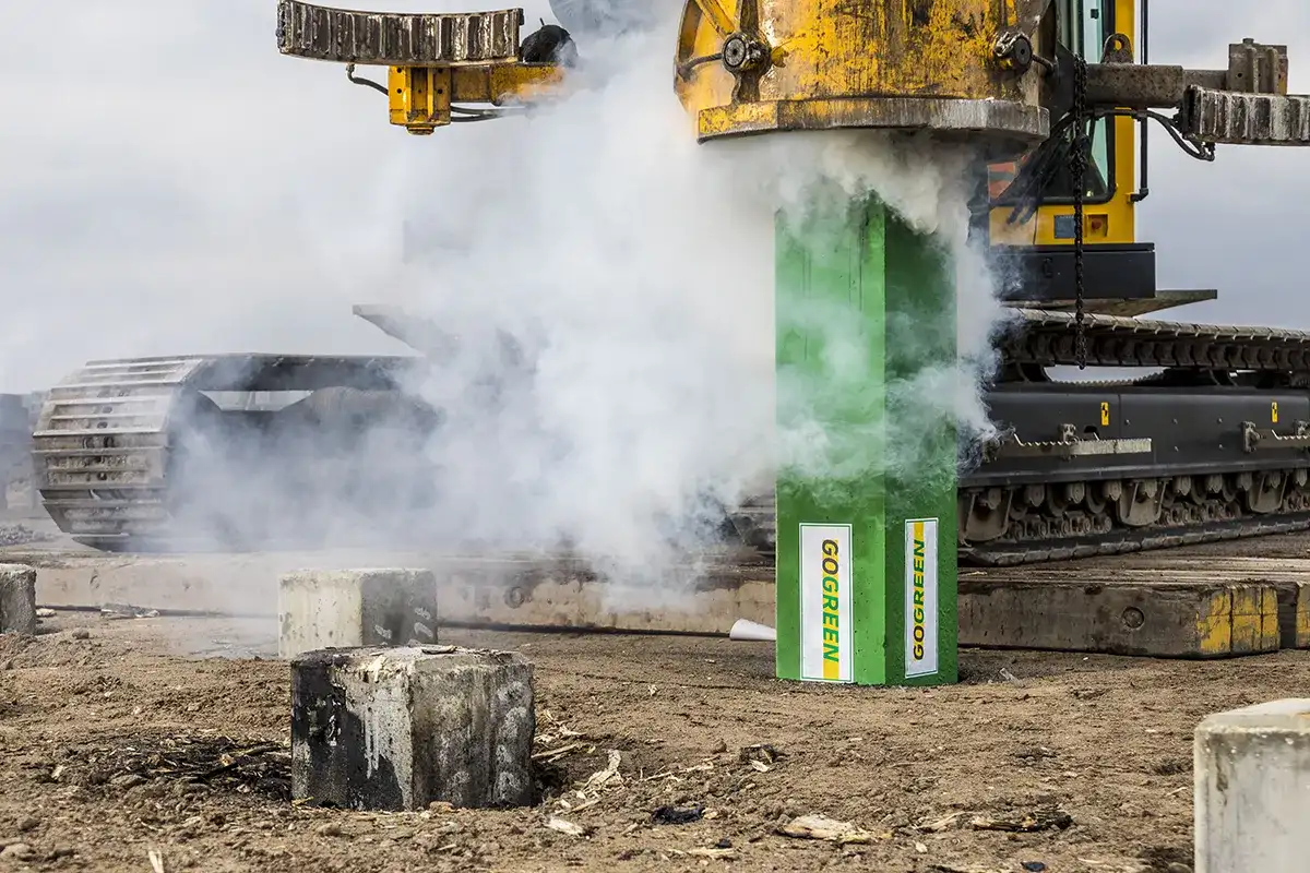 Een grote, gele industriële boormachine werkt op een bouwplaats. De machine stoot witte rook uit terwijl deze werkt op een groene cilinder met het opschrift 'GOCREEN'. Op de grond rond de machines zijn betonnen palen en vuil zichtbaar. Op de achtergrond is de lucht bewolkt.