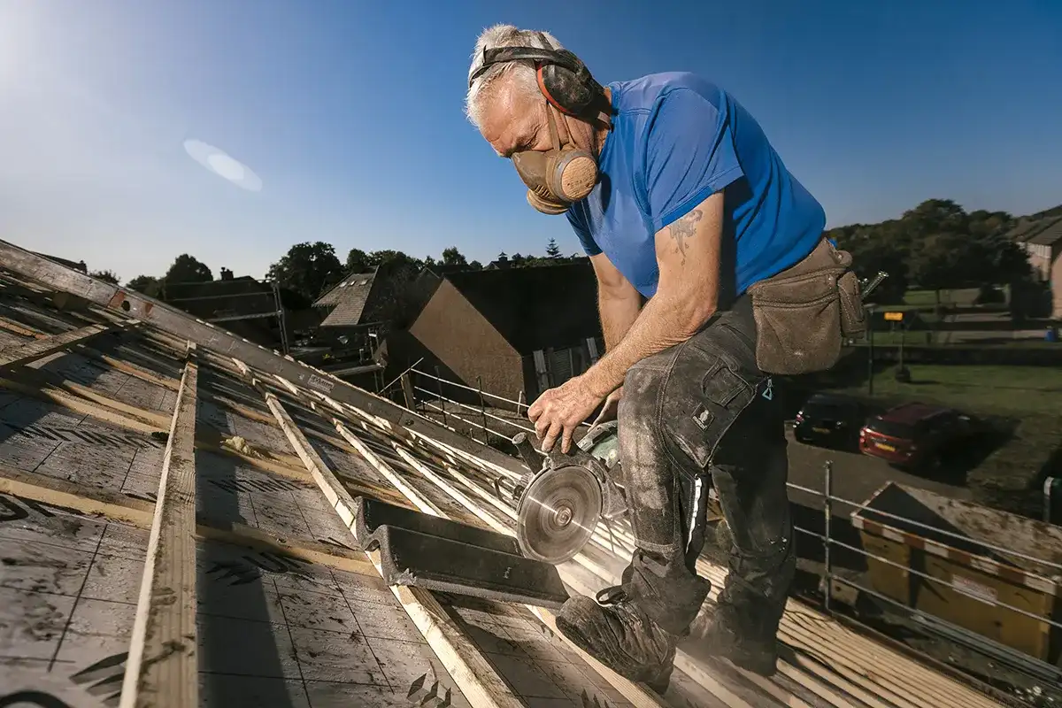 Een oudere man met gehoorbescherming, veiligheidsbril, een stofmasker, een blauw shirt en een bruine gereedschapsriem gebruikt een cirkelzaag op een houten dakframe. De lucht is helder en blauw en op de achtergrond zijn huizen en bomen zichtbaar.
