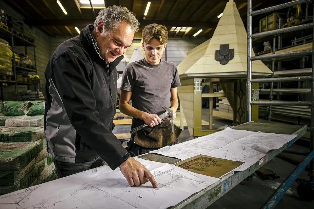 Twee mannen staan in een werkplaats naast een grote tafel vol bouwplannen. De oudere man wijst naar de plannen, glimlacht en draagt een zwart-grijs jasje. De jongere man, met een hamer in zijn hand, kijkt aandachtig toe, gekleed in een grijs t-shirt. Op de achtergrond staat een klein houten bouwwerk.