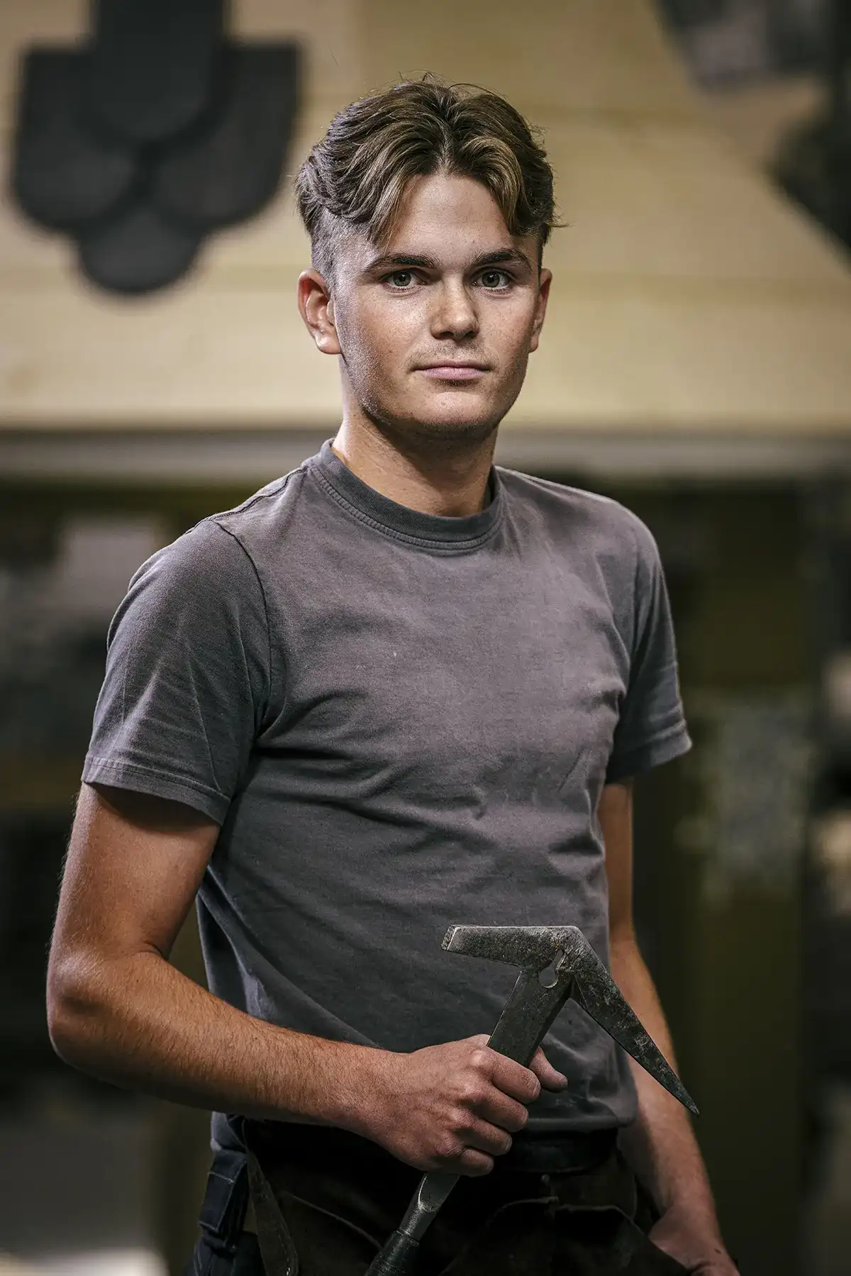 Een jonge man staat zelfverzekerd binnenshuis, met een smidse hamer in zijn hand. Hij heeft kort, licht golvend haar met een scheiding in het midden, een flauwe glimlach en draagt een grijs T-shirt. De achtergrond is wazig, maar toont werkplaatsdetails, waaronder een smederij en gereedschap, waardoor de setting van een smid wordt benadrukt.
