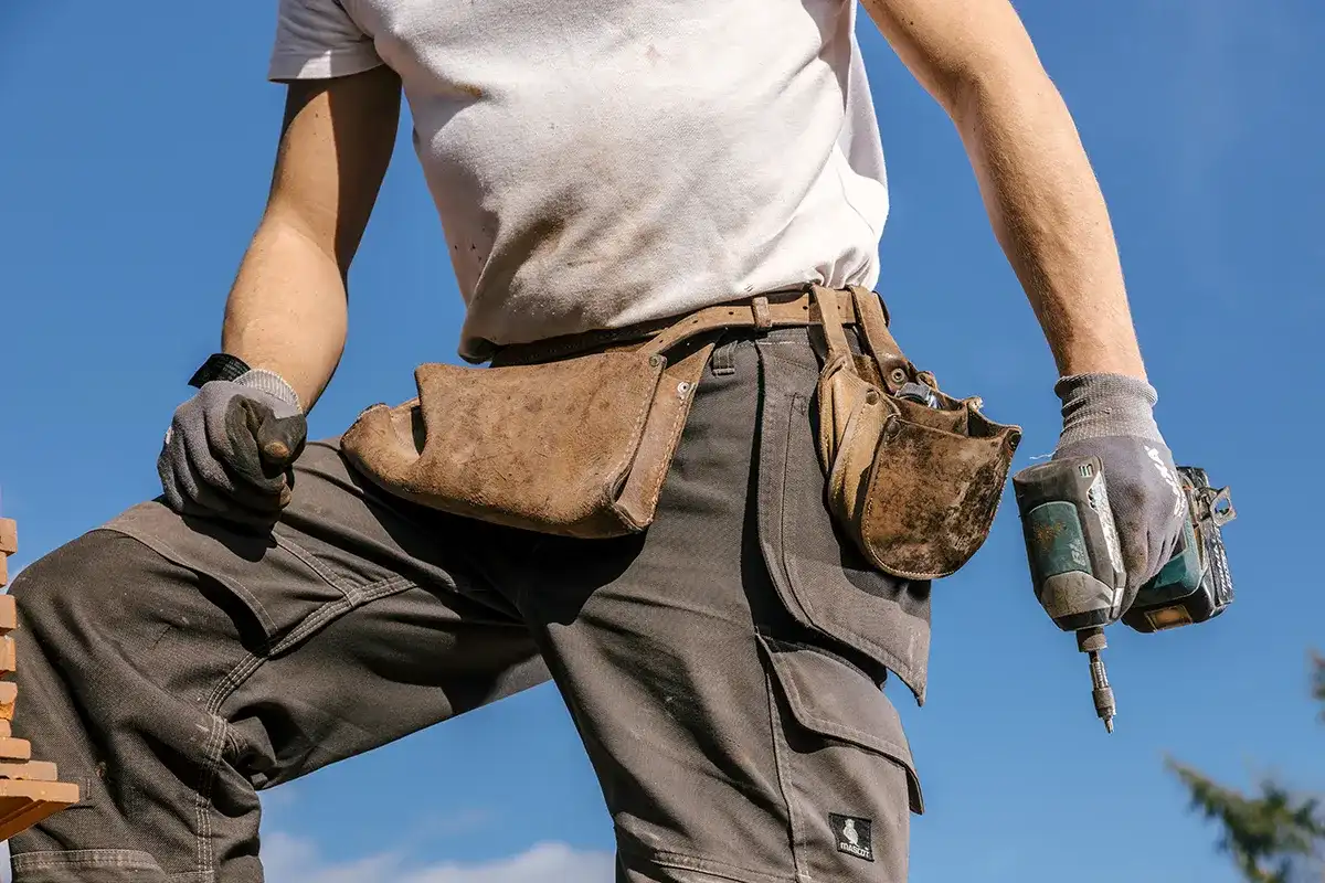 Een persoon die een wit T-shirt, een grijze werkbroek en handschoenen draagt, gebruikt een boormachine. Ze hebben een lichtbruine leren gereedschapsriem met meerdere zakjes om hun middel, gevuld met gereedschap. De achtergrond is een helderblauwe lucht, wat aangeeft dat ze buiten aan het werk zijn.