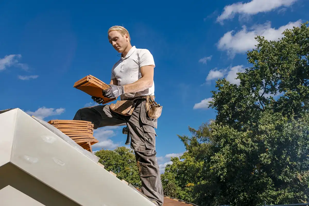 Een persoon, gekleed in een wit poloshirt, een grijze cargobroek en een gereedschapsriem, staat op een dak. Ze plaatsen dakpannen van klei op een stapel. De achtergrond heeft een helderblauwe lucht met verspreide witte wolken en groene bomen. De persoon lijkt gefocust op zijn werk.