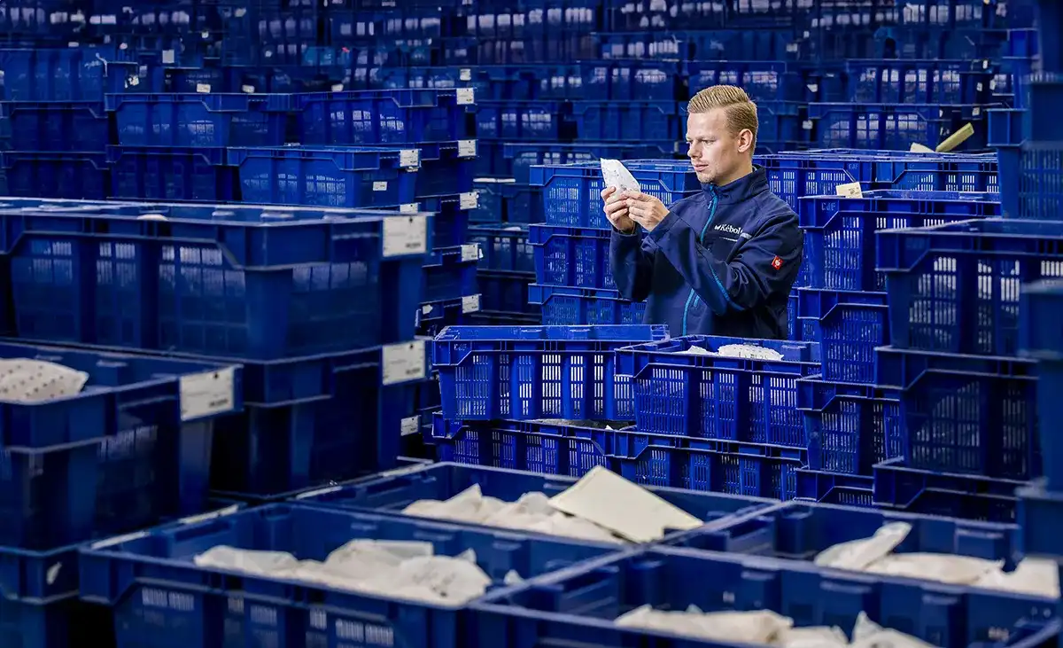 Een arbeider in een blauw uniform inspecteert het papierwerk, omringd door talloze gestapelde blauwe plastic kratten in een magazijn. De kratten, gevuld met diverse papieren en documenten, creëren een doolhofachtige omgeving met een industriële sfeer. De man lijkt geconcentreerd en leest aandachtig het papierwerk.
