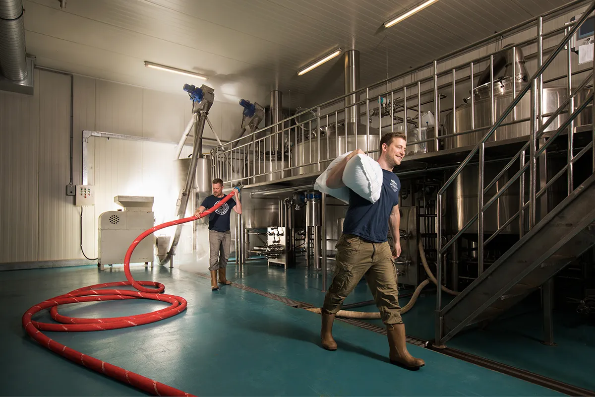 Twee mannen in een brouwerij aan het werk. Eén man aan de rechterkant draagt een bulkzak over zijn schouder, terwijl een andere man aan de linkerkant een lange rode slang hanteert. De kamer is ruim en heeft blauwgroen vloeren, roestvrijstalen tanks en industriële apparatuur die doet denken aan brouwactiviteiten. Beiden dragen casual werkkleding.