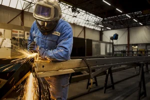 Een lasser in blauw werkpak en lashelm slijpt vonkend staalwerk op een bok in een grote industriële hal, documentair en rauw belicht