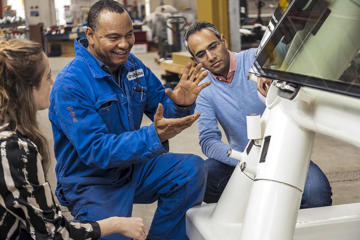 Drie mensen zitten rond een industrieel apparaat. De persoon links, een vrouw met lang haar in een zebragestreept topje, luistert aandachtig. De man in het midden, gekleed in een blauwe jumpsuit, legt met expressieve handgebaren iets uit. De meest rechtse man met bril en blauwe trui kijkt aandachtig toe.