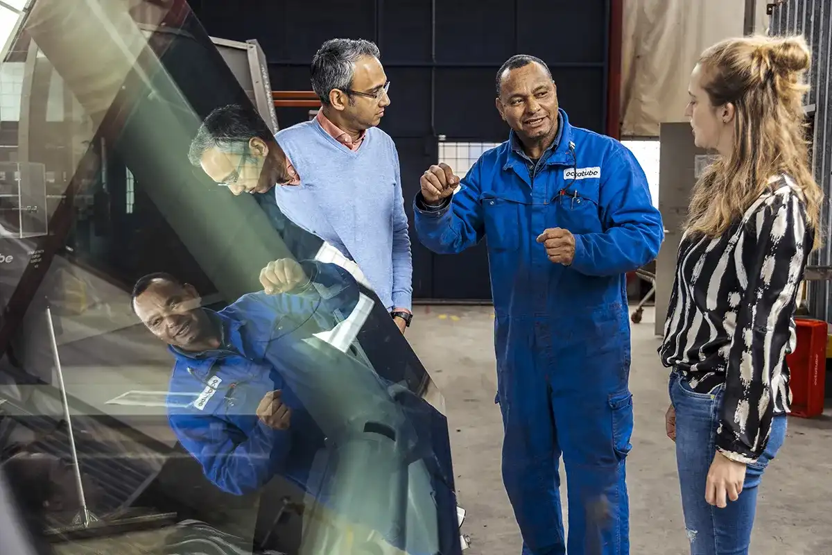 Drie mensen staan binnen en praten. De persoon in het midden, gekleed in een blauwe jumpsuit, gebaart terwijl hij praat met de andere twee, die nonchalant gekleed zijn. Ze bevinden zich in de buurt van grote glazen panelen, die het tafereel weerspiegelen. De omgeving lijkt industrieel.