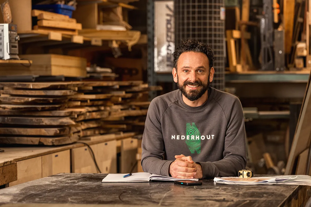 Een man in een grijze Nederhout-sweater zit glimlachend aan een werktafel in een houtbewerkingswerkplaats, omringd door gestapelde houten planken, warm en authentiek belicht.