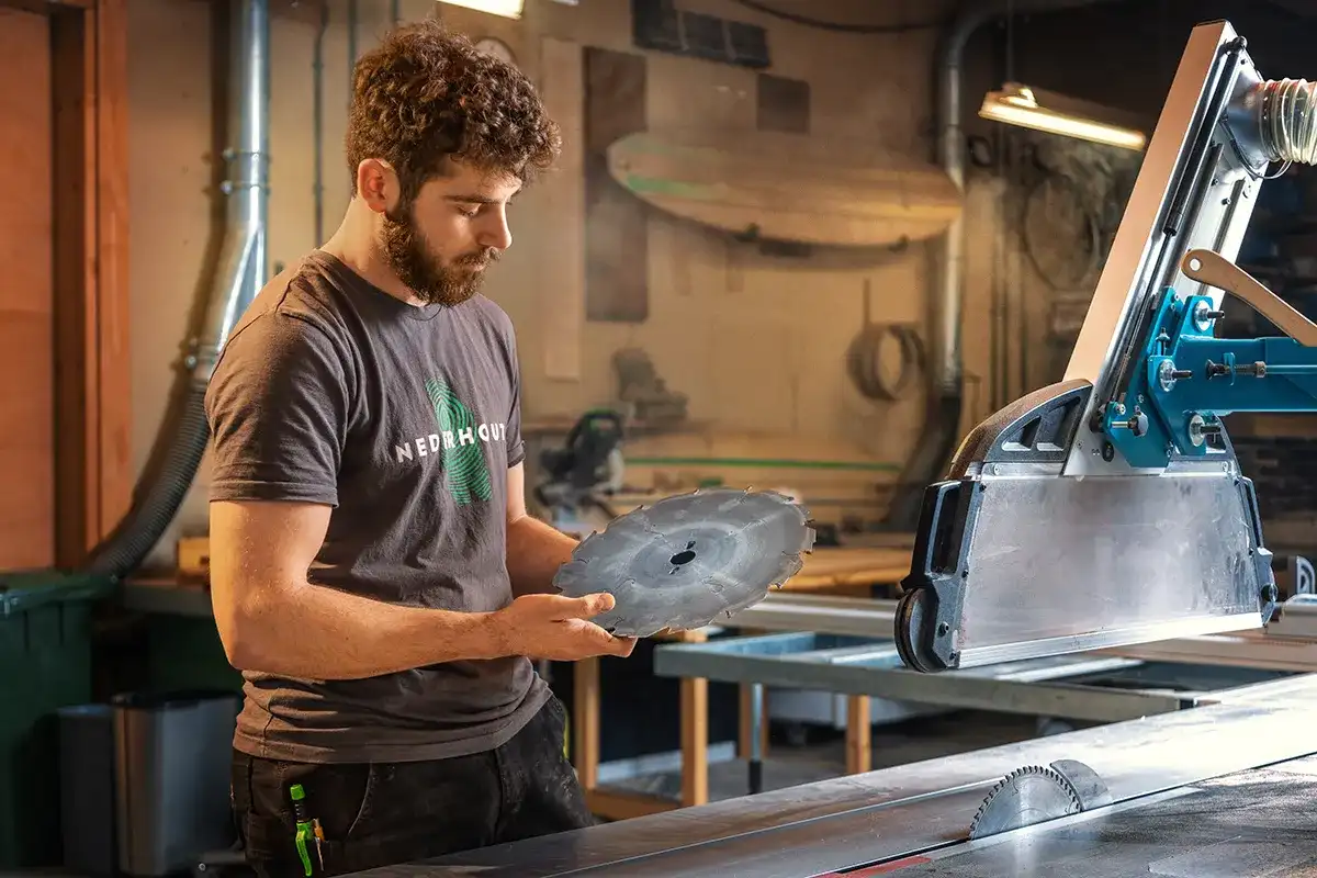 Een man met een bruine baard en krullend haar staat in een werkplaats met een groot cirkelzaagblad in zijn hand. Hij lijkt gefocust terwijl hij het mes onderzoekt. Hij draagt een grijs T-shirt met een gedeeltelijk verborgen logo. De werkplaats, vermeld op onze homepage, is uitgerust met diverse gereedschappen en machines, waaronder een zaagtafel.