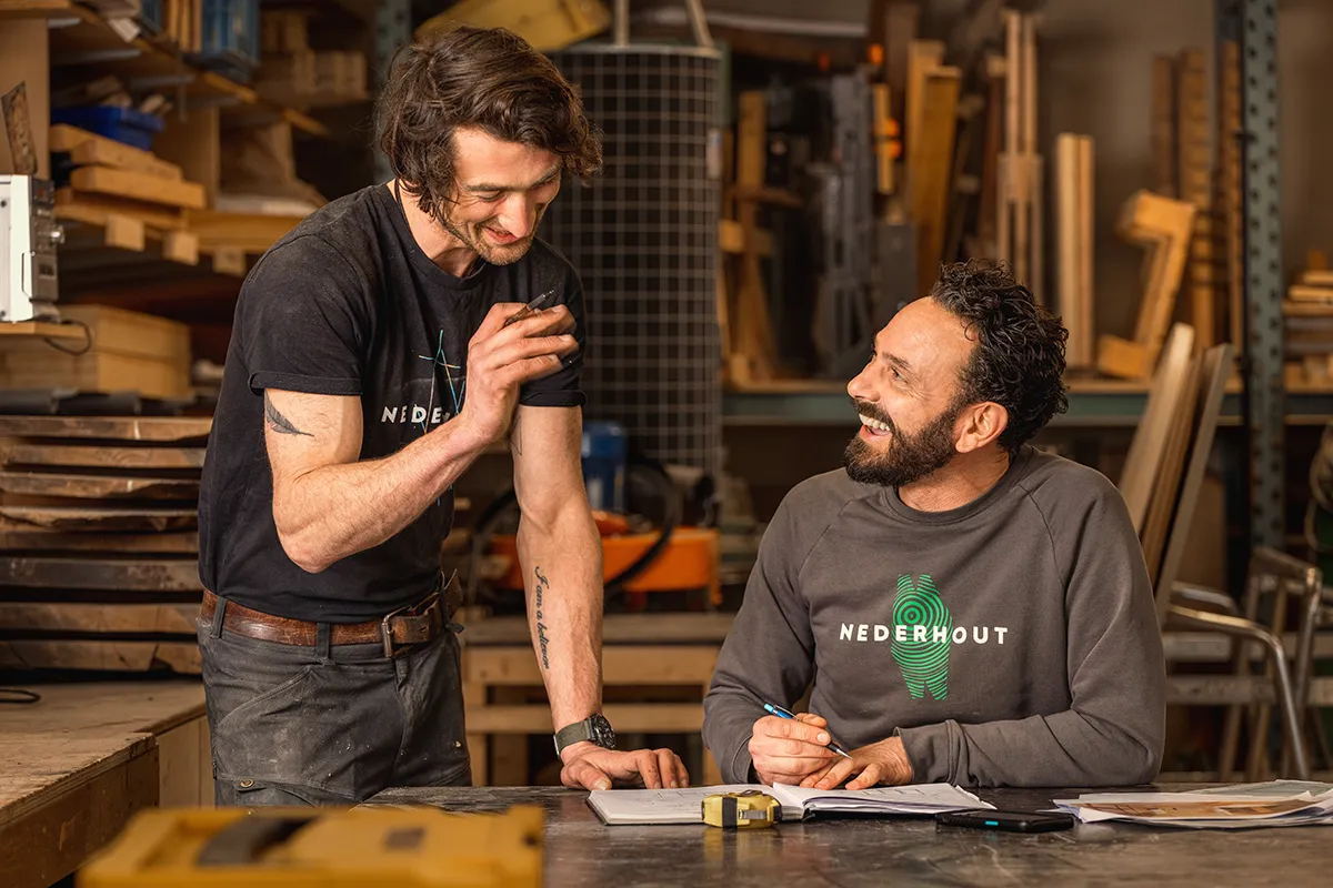 Twee mannen in een houtwerkplaats. Eén, staande, houdt nonchalant een telefoon vast en lijkt te praten of te lachen. De ander, zittend, glimlacht hartelijk terwijl hij een pen vasthoudt. Beiden dragen donkere shirts met het Nederhout-logo. De achtergrond gevuld met houten materialen, gereedschap en planken ademt een gezellige huiselijke sfeer uit.