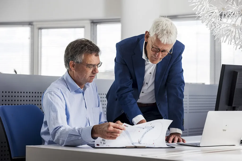 Twee oudere mannen in zakelijke kleding werken samen in een modern kantoor. De één zit en wijst naar een papier met diagrammen, terwijl de ander zich voorover buigt om te bespreken. Op het bureau staan een computer, een open laptop en documenten. Grote ramen op de achtergrond laten voldoende natuurlijk licht binnen, perfect voor bedrijfsfotografie.