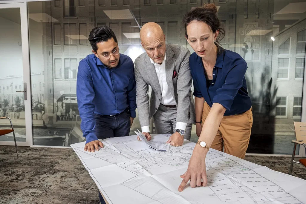 Drie individuen staan rond een tafel bedekt met architectonische blauwdrukken in een kantooromgeving, perfect voor bedrijfsfotografie. De persoon links draagt een blauw shirt en een bril. In het midden onderzoekt een kaal individu in een grijs pak de plannen. Rechts wijst iemand in een blauwe blouse naar de blauwdrukken.