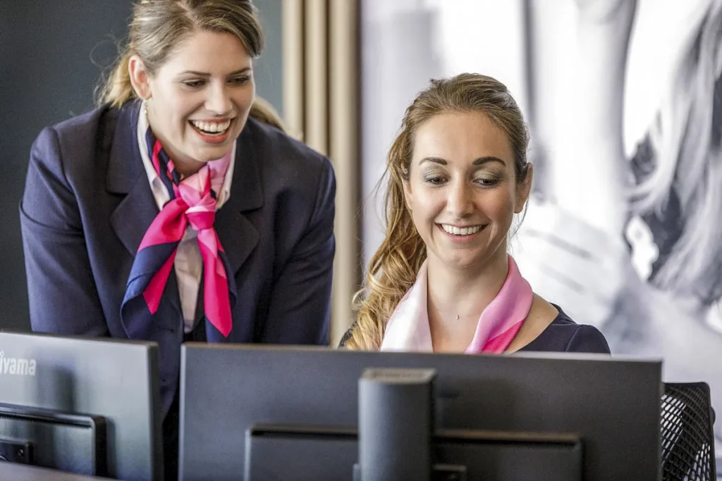 Twee vrouwen in professionele kleding, met bijpassende roze sjaals, zijn met elkaar bezig terwijl ze op computerstations werken. De ene vrouw zit en lacht naar het scherm, terwijl de andere, die naast haar staat, ook lacht en naar het scherm kijkt: een perfect moment voor bedrijfsfotografie.