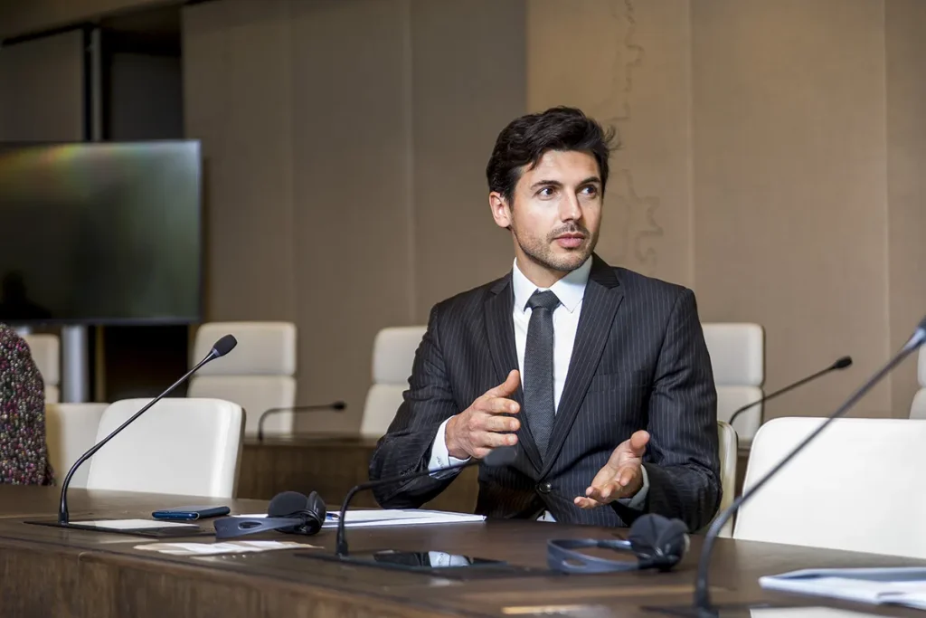 Een man in een donker krijtstreeppak en stropdas zit aan een vergadertafel en gebaart met zijn handen terwijl hij spreekt. Op de tafel zijn microfoons en koptelefoons geplaatst. De beige muren en lege witte stoelen op de achtergrond weerspiegelen de professionele setting, ideaal voor bedrijfsfotografie.