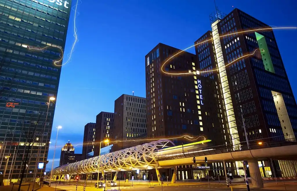 Een stadsgezicht bij nacht met verlichte moderne hoogbouw, waaronder een hoogbouw met een zichtbaar bord 'HOTEL'. Een webachtige voetgangersbrug overspant de straat beneden. Lichtsporen van verkeer accentueren het tafereel en leggen levendige bedrijfsfotografie vast tegen een diepblauwe lucht op de achtergrond.