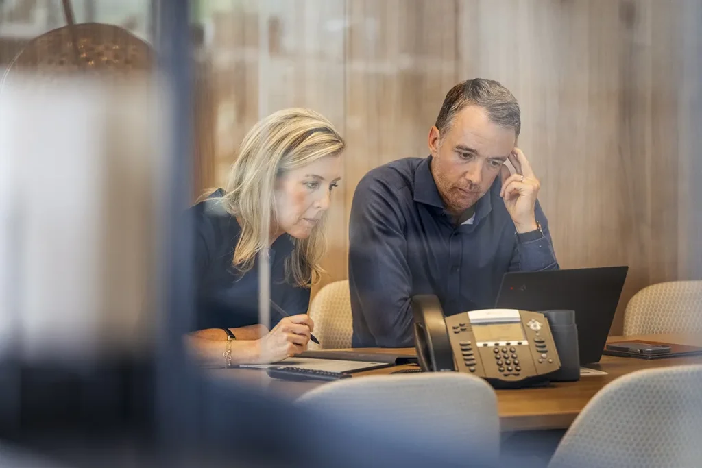 Een man en een vrouw zitten aan een houten tafel, intens gefocust op een laptop. De man, gekleed in een donkerblauw shirt, raakt zijn slaap aan, terwijl de vrouw, eveneens in donkere kleding, naar het laptopscherm naast hem kijkt. Op de tafel staat een conferentietelefoon tegen een achtergrond van houten wandpanelen: perfect voor bedrijfsfotografie.