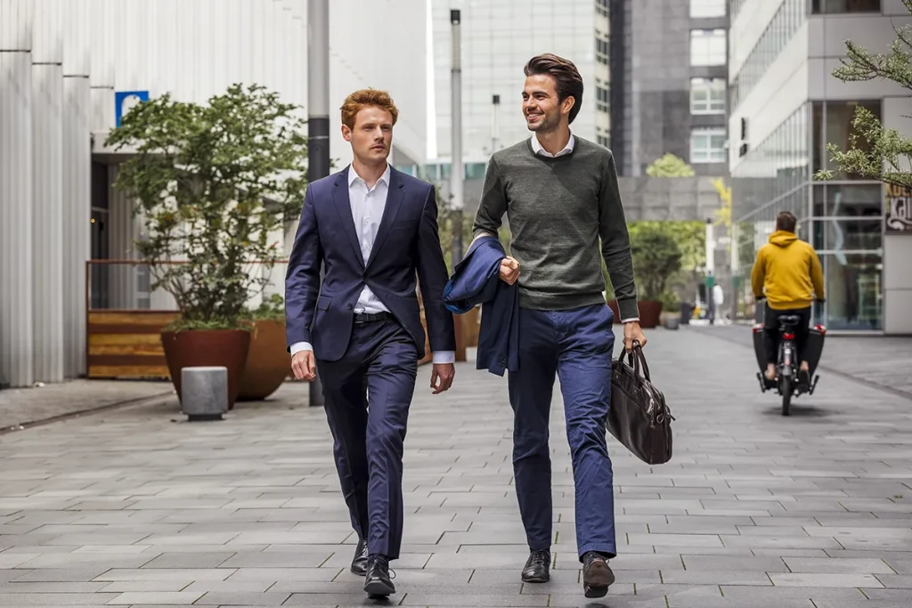 Twee mannen lopen door een moderne stadsstraat. De een, met rood haar, draagt een blauw pak en een wit overhemd, terwijl de ander, met donker haar, een groene trui over een blauw overhemd en een pantalon draagt, terwijl hij een jasje en een aktetas vasthoudt. Op de achtergrond, die de essentie van bedrijfsfotografie vastlegt, fietst een persoon in een gele top.