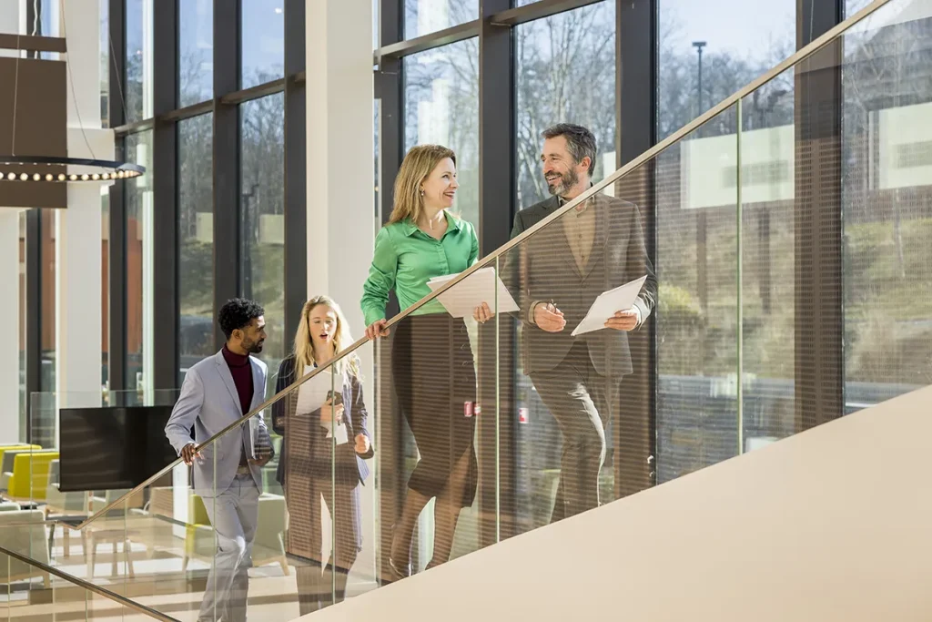 Vier zakenmensen lopen een trap met glazen wanden op. Vooraan zijn een man in pak en een vrouw in een groene blouse, beiden met documenten in de hand, in gesprek. Achter hen stijgen ook nog een man en een vrouw op. Helder natuurlijk licht vult de moderne, open kantoorruimte: een ideale scène voor bedrijfsfotografie.