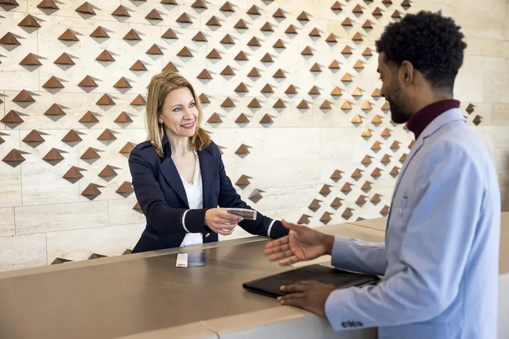 Een vrouw bij de receptie van een hotel overhandigt een sleutelkaart aan een man aan de overkant van de balie. De vrouw heeft blond haar en draagt een marineblauwe blazer, terwijl de man, met krullend haar en baard, een lichtblauw jasje draagt. De achtergrond is voorzien van een decoratieve muur met driehoekige patronen, perfect voor bedrijfsfotografie.