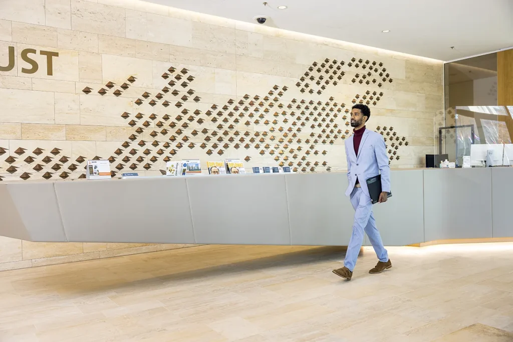 Een man in een lichtblauw pak, kastanjebruine coltrui en lichtbruine schoenen loopt door een moderne kantoorlobby met lichtgekleurde tegelvloeren en een beige, gestructureerde muur met metalen accenten. Hij heeft een zwarte map bij zich. Op de receptiebalie zijn diverse spullen uitgestald en aan de rechterkant zit glas: een perfecte setting voor bedrijfsfotografie.