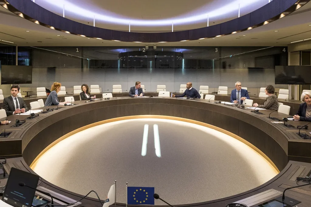 Een groep mensen in zakelijke kleding zit rond een grote, ronde vergadertafel met microfoons en documenten. De kamer heeft een modern design en zachte, warme verlichting, perfect voor bedrijfsfotografie. Op de tafel liggen een EU-vlag en een andere vlag. Glazen wanden op de achtergrond scheiden de kamer van andere ruimtes.