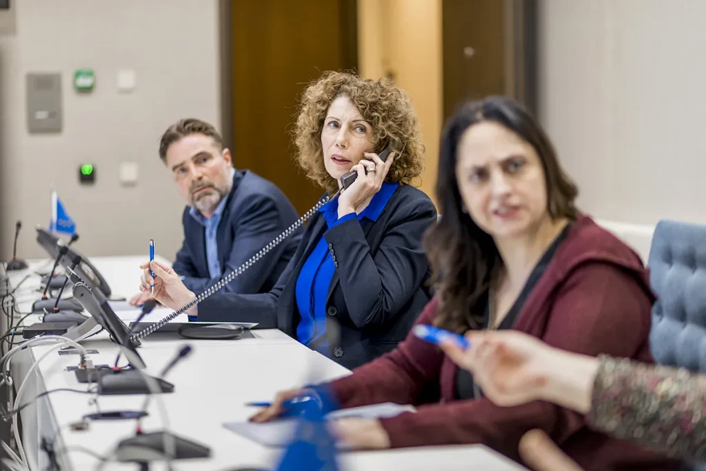 Drie personen zitten aan een lange tafel met elkaar te discussiëren. De vrouw in het midden, met krullend haar en gekleed in een blauw topje en een zwarte blazer, is aan het telefoneren. De man links van haar en de vrouw rechts van haar zijn verloofd, terwijl de vrouw rechts naar iemand buiten beeld kijkt – perfect voor bedrijfsfotografie.