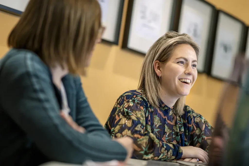 In een helder verlichte kamer zitten twee vrouwen aan een tafel. De vrouw rechts, met blond haar op schouderlengte, glimlacht in een blouse met bloemenmotief. De vrouw links, met halflang bruin haar en een bril, draagt een groen topje. Ingelijste foto's sieren de gele muur erachter en geven de essentie van bedrijfsfotografie weer.
