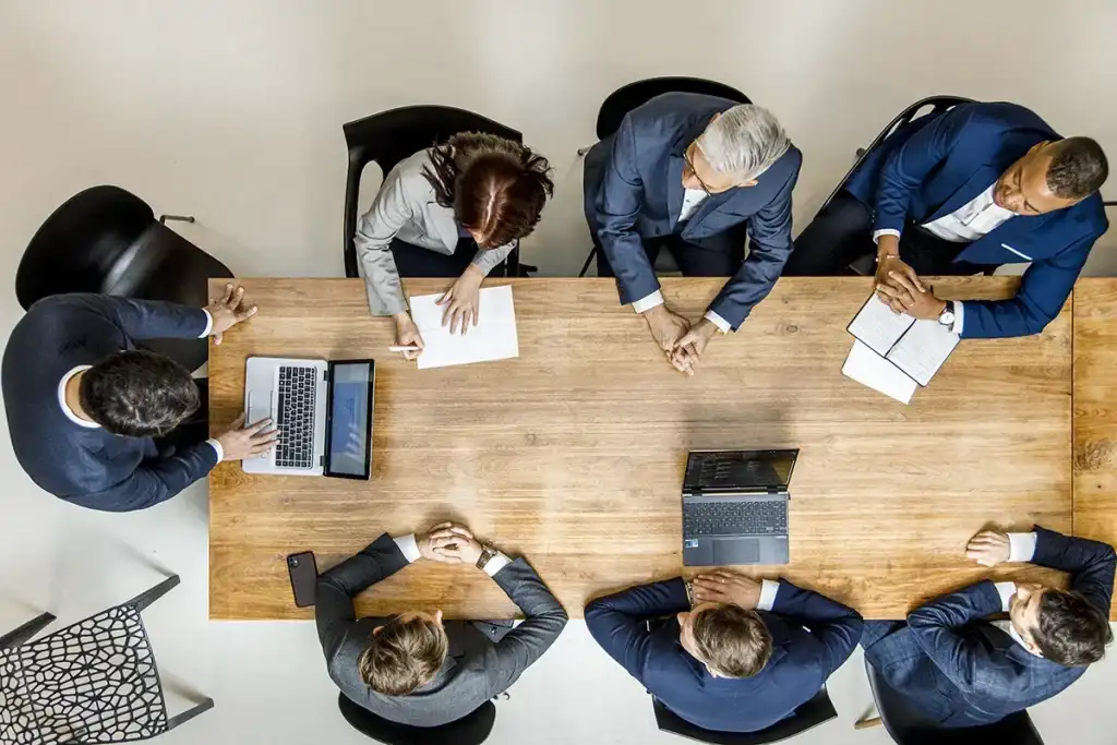 Luchtfoto van een zakelijke bijeenkomst met zes personen rond een houten tafel. Aan elke kant staan drie mensen, waarvan er één op een laptop typt, twee aantekeningen maken en de rest in discussie is. Papieren en nog een laptop liggen op tafel. Deze scène legt professionele bedrijfsfotografie op zijn best vast.
