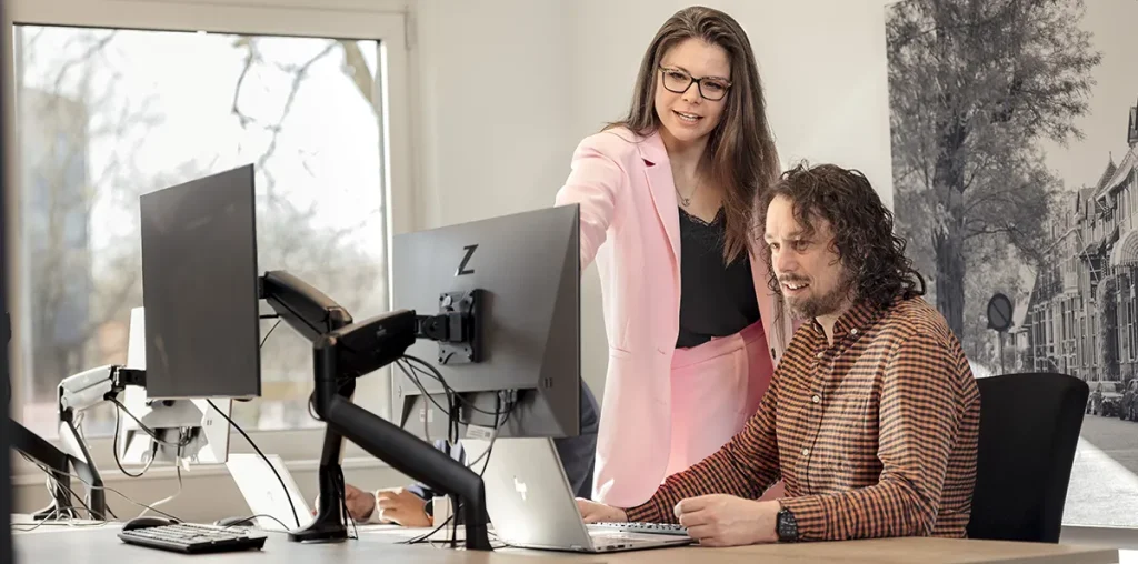 In een kantoor met grote ramen en meerdere monitoren op de bureaus staat een vrouw in een roze pak en wijst naar een computerscherm, terwijl een man met krullend haar en een geruit overhemd naar zijn laptop zit te kijken. Ze lijken nauw samen te werken, wat de essentie van bedrijfsfotografie weerspiegelt.