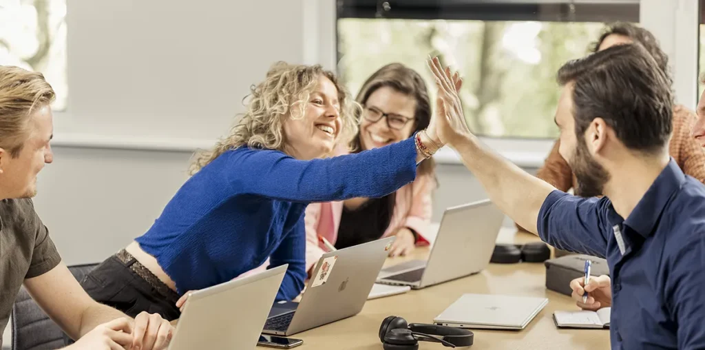 Een groep van vijf collega's in een vergaderruimte viert feest met een high five. De vrouw in een blauwe trui reikt over de tafel heen en geeft een high five aan een bebaarde man in een grijs overhemd. Ze worden omringd door laptops en notebooks, terwijl natuurlijk licht door het heldere raam naar binnen valt, wat perfecte bedrijfsfotografie oplevert.