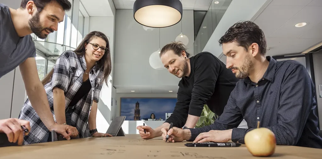Vier mensen werken samen rond een grote tafel in een modern kantoor, perfect vastgelegd in dit bedrijfsfotografiemoment. Twee mannen en een vrouw staan terwijl een andere man, zittend, met stiften op papier tekent. De vrouw houdt een laptop vast. Erboven hangt een grote ronde lamp en op de rand van de tafel staat een appel tegen de achtergrond van een glazen wand.