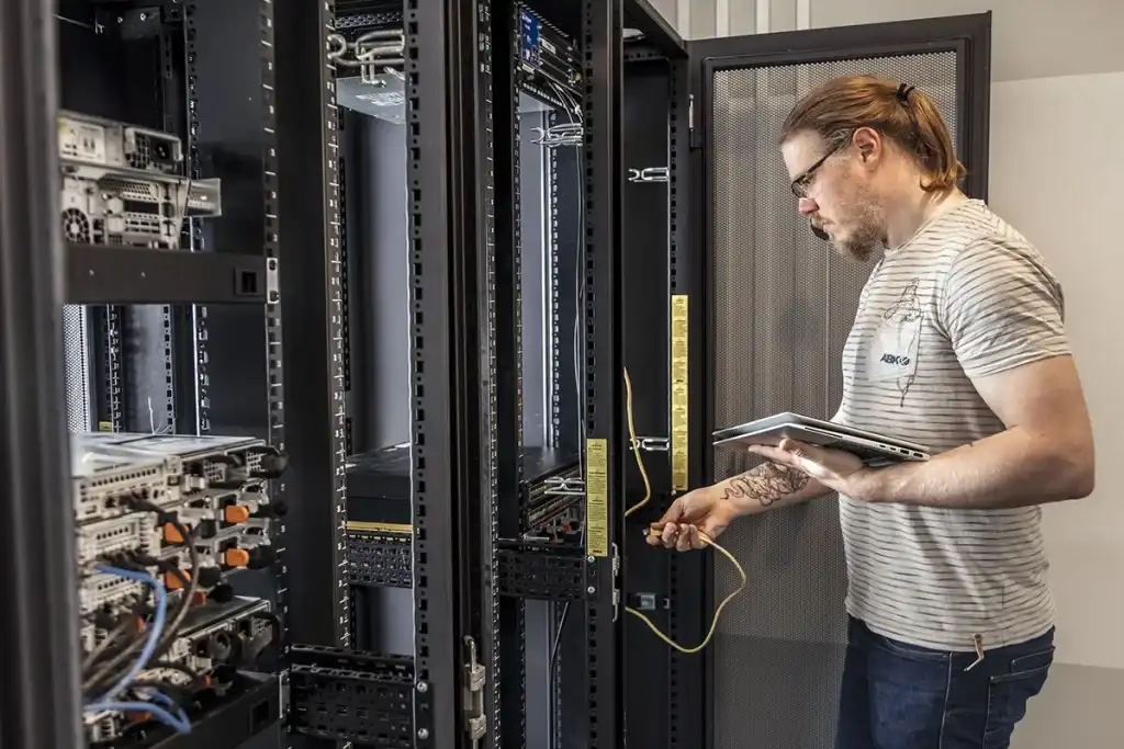 In een serverruimte houdt een man met achterovergetrokken haar en een bril, gekleed in een gestreept T-shirt en een spijkerbroek, een gele netwerkkabel en een tablet vast. Hij concentreert zich op het onderzoeken van een van de serverrekken vol elektronische apparatuur en kabels. Deze scène vat de essentie van bedrijfsfotografie samen.