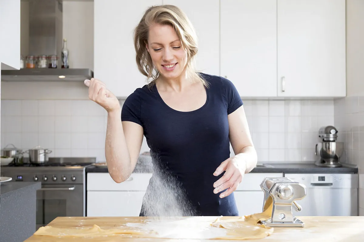 Een vrouw in een marineblauw overhemd glimlacht terwijl ze bloem op een houten aanrecht strooit. Ze bevindt zich in een moderne keuken met witte kasten en roestvrijstalen apparaten. Voor haar op het aanrecht staan vers gerold pastadeeg en een metalen pastamachine.