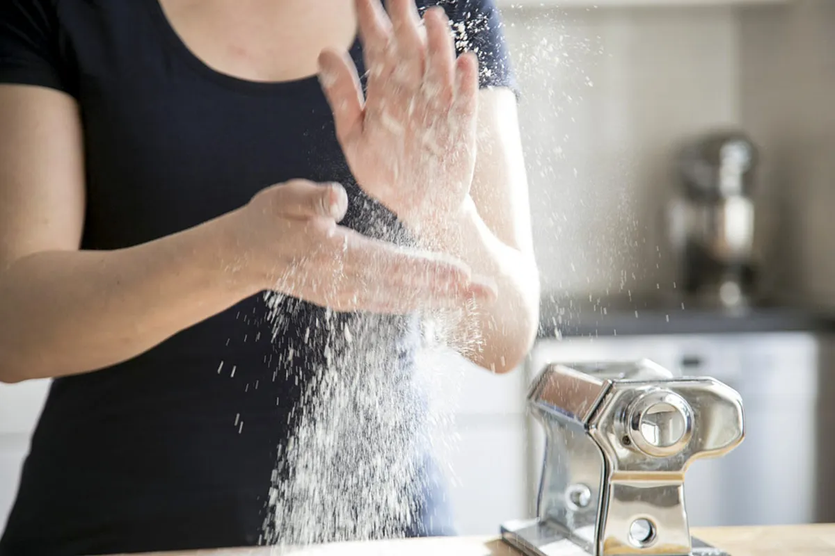 Een persoon die een zwart shirt draagt, stoft bloem van zijn handen over een houten oppervlak. Een onafgemaakte pastamachine staat vlakbij op het aanrecht. De achtergrond toont een wazige keukenomgeving met lichtgekleurde kasten. De scène weerspiegelt de voorbereiding voor het maken van pasta.